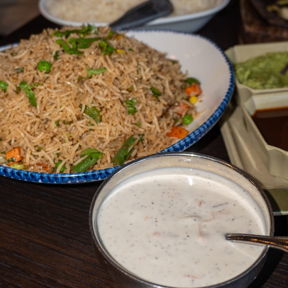 A bowl of raita with a spoon, next to a plate of vegetable fried rice on a wooden table.