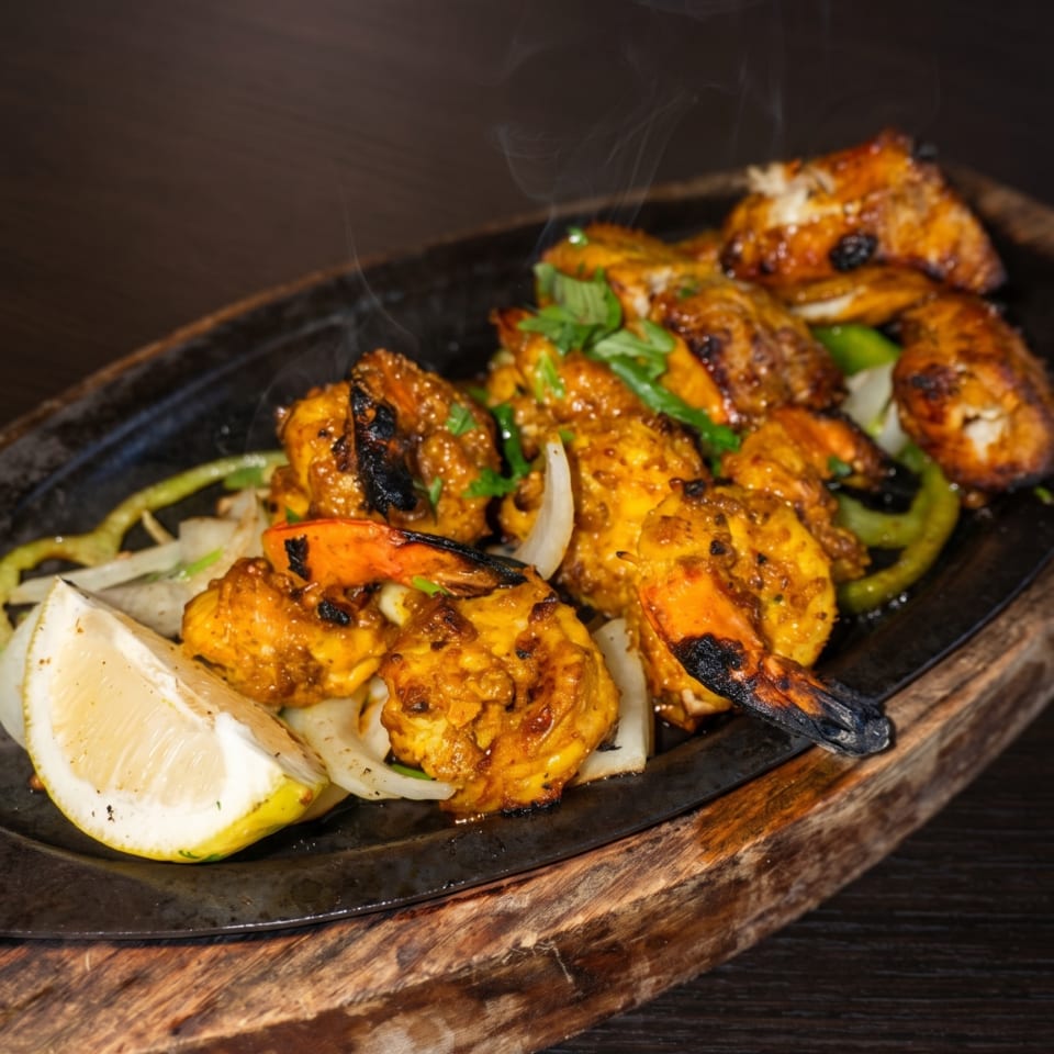 Sizzling shrimp and chicken tikka with onions, peppers, and lemon on a dark wooden board.