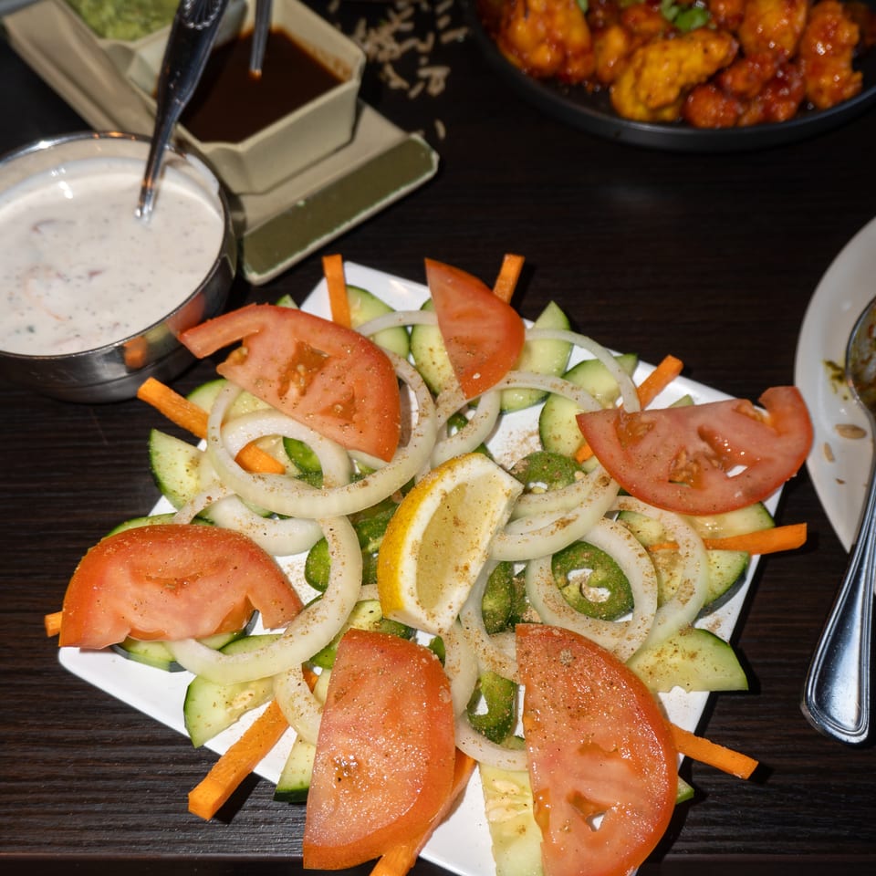 Fresh salad with sliced tomatoes, cucumbers, onions, jalapeños, and a lemon wedge, next to sauces and chicken.