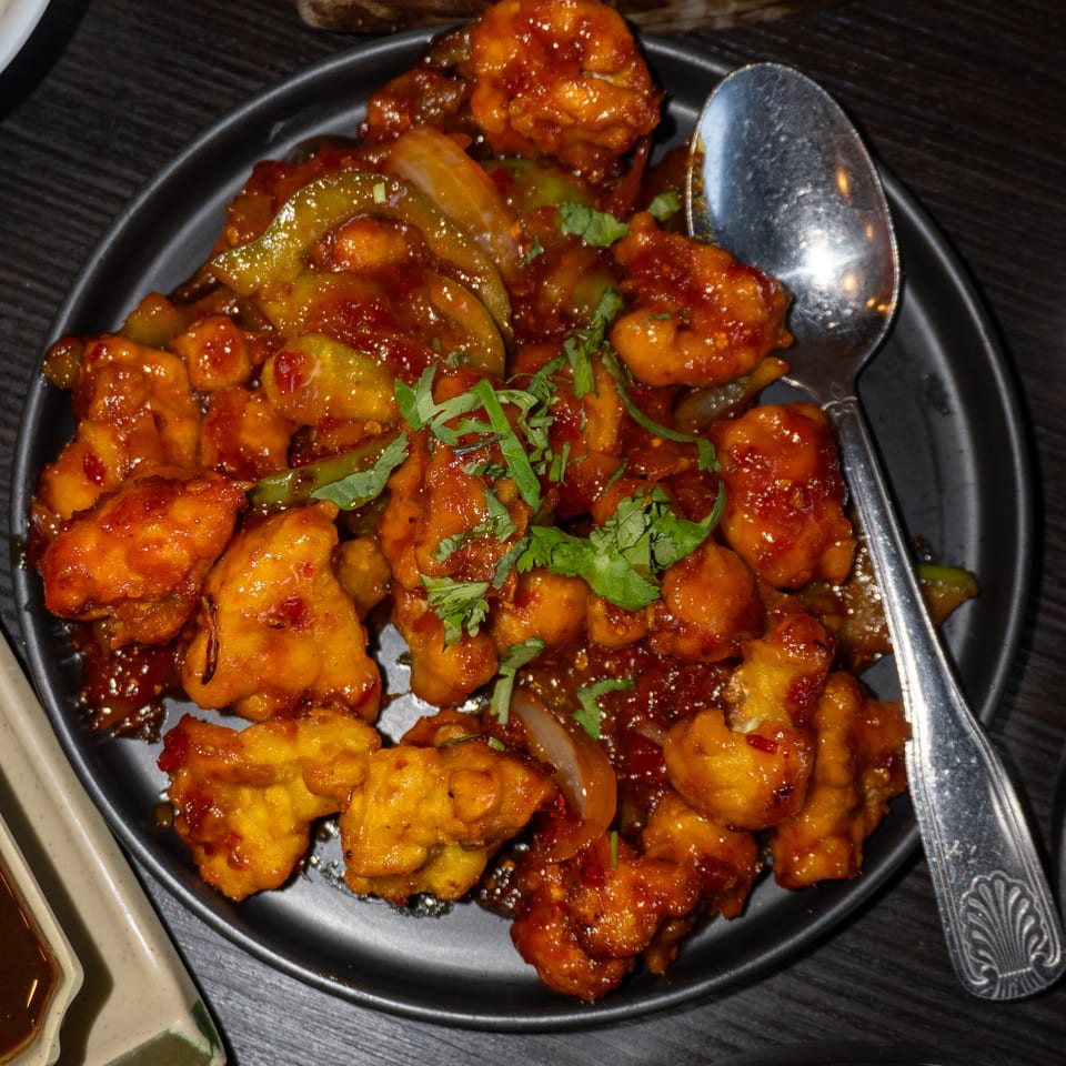 A black plate of orange-glazed chili chicken with green peppers and cilantro, with a spoon.